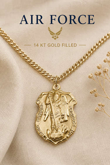 A gold Air Force pendant necklace displayed on a beige fabric background with decorative dried flowers. The image includes text reading 'AIR FORCE' and '14 KT GOLD FILLED' with a winged emblem above.